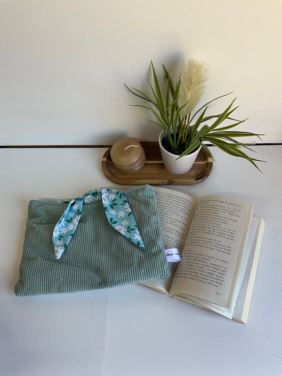 Pochette à livre horizontale en velours côtelé vert sauge, doublure marguerites – posée sur un livre ouvert, cousue main par Sidonie Créa