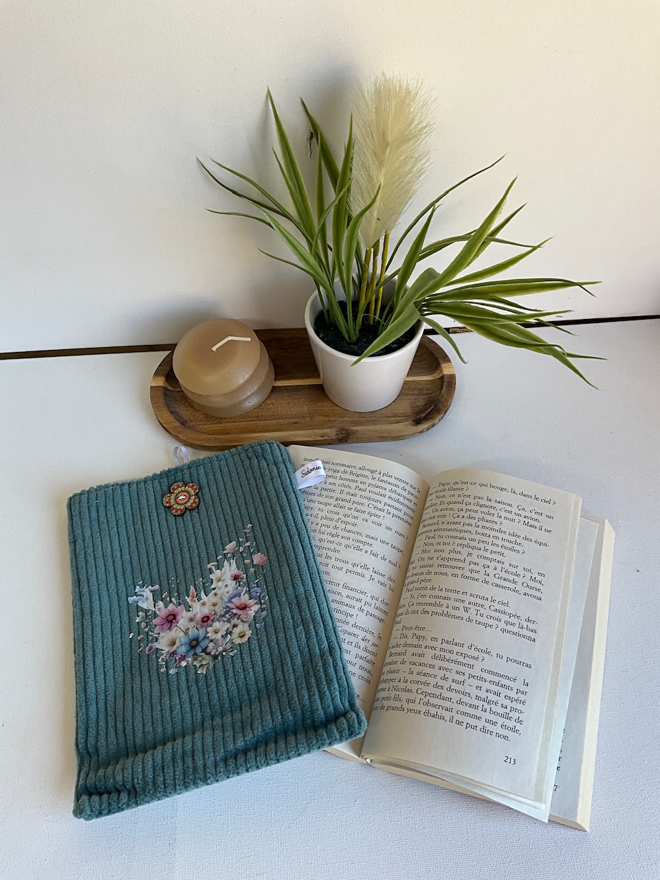 Pochette à livre verticale en velours côtelé bleu canard, appliqué couronne de fleurs – posée sur un livre ouvert, cousue main par Sidonie Créa