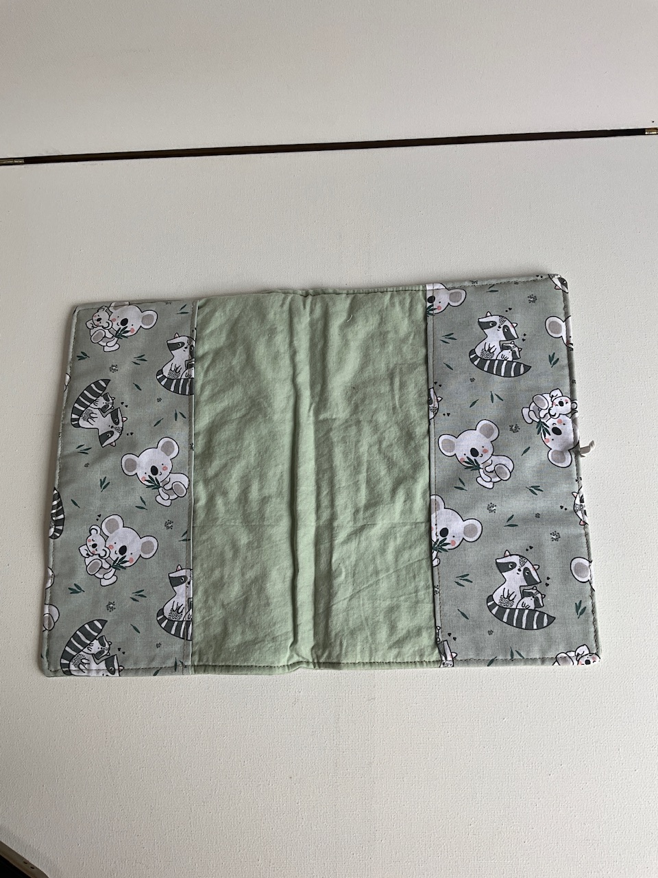 Protège carnet de santé bébé ouvert, motif koalas et ratons laveurs, deux rabats latéraux et panneau central vert sauge dans lesquels se glisse la couverture du carnet
