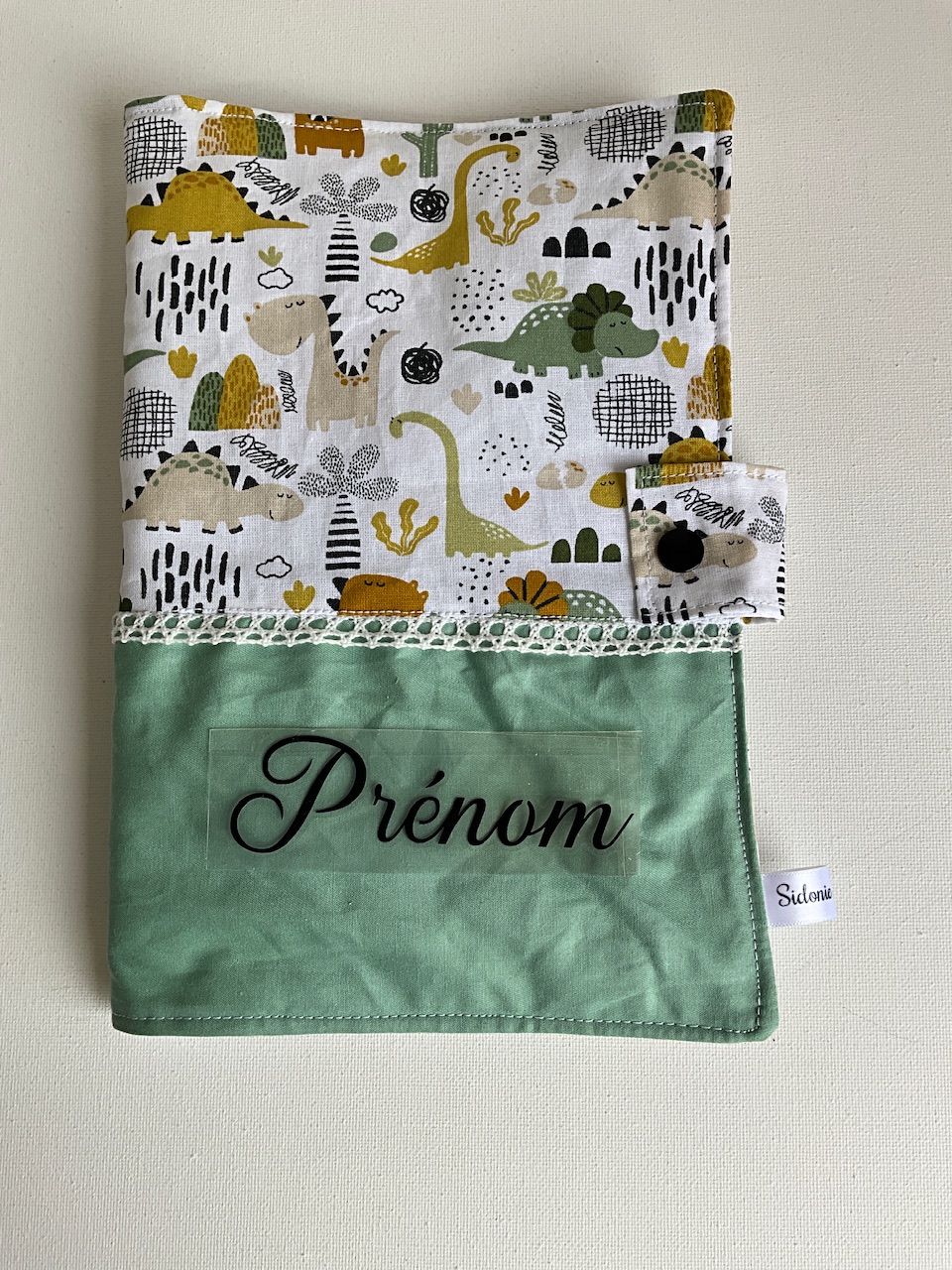 Protège carnet de santé personnalisable en coton dinosaures ocre et kaki fond écru – vu de face avec le prénom floqué, cousu main par Sidonie Créa